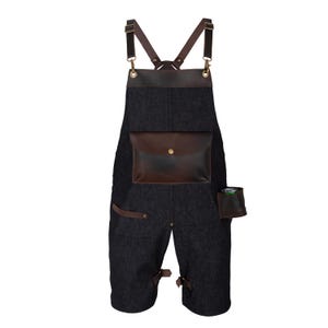 Könnte beinhalten: Dunkelgraue Denim-Schürze mit braunen Lederakzenten. Sie verfügt über eine große Fronttasche, eine kleinere Seitentasche und verstellbare Lederriemen. Die Schürze hat einen Knopfverschluss an der Vordertasche und Schnallen an den Beinen.