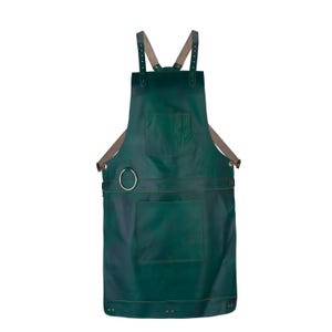 May include: A dark green leather apron featuring adjustable beige shoulder straps, a chest pocket, a large lower pocket, and a silver-toned metal ring attached to the side for hanging tools. The apron is secured with metal rivets.