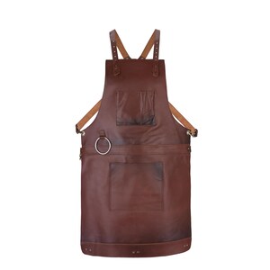 May include: A dark brown leather apron featuring adjustable tan shoulder straps, a chest pocket, a large lower pocket, and a silver-toned metal ring attached to the side for utility. The apron is secured with metal rivets and hardware.