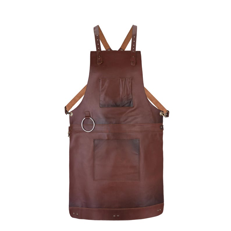 May include: A dark brown leather apron featuring adjustable tan shoulder straps, a chest pocket, a large lower pocket, and a silver-toned metal ring attached to the side for utility. The apron is secured with metal rivets and hardware.