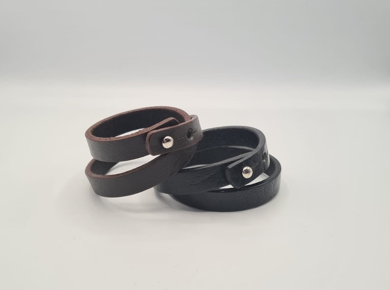May include: Two leather bracelets, one brown and one black, with silver metal accents. The bracelets are wrapped around each other.