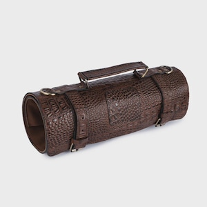 May include: A cylindrical brown case with a textured crocodile skin pattern. The case features a top handle, two adjustable buckled straps, and metallic D-rings on each end. The interior is lined with a smooth brown material, showing an open end.