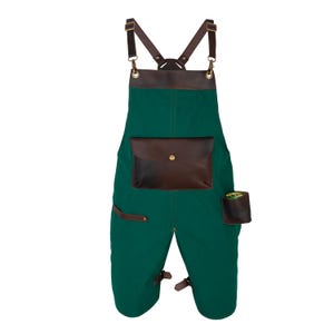 Könnte beinhalten: Grüne Garten-Latzhose aus Canvas mit braunen Lederdetails. Mit einer großen Fronttasche, einer kleinen Seitentasche und verstellbaren braunen Lederriemen. Die Latzhose hat einen Latz-Stil und bedeckt die Beine.