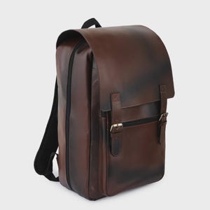 Puede incluir: Una mochila de cuero marrón oscuro con un acabado liso y un sutil efecto degradado. La bolsa cuenta con una solapa superior grande asegurada por dos correas verticales con hebillas de metal plateado, un bolsillo frontal con cremallera y correas negras.