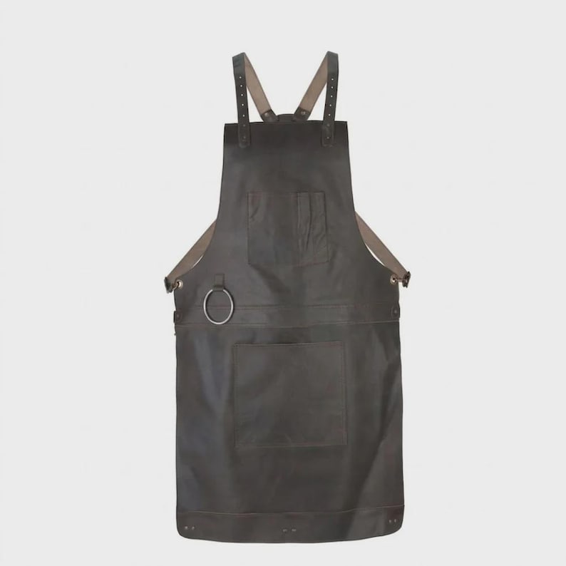 May include: A dark brown leather apron featuring adjustable shoulder straps with metal buckles, a chest pocket, a large lower pocket, and a circular metal ring attached to the side for hanging tools. The apron has a smooth, matte finish.