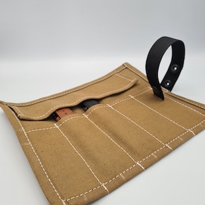 May include: A tan canvas watch roll with white stitching and a black strap with two snap buttons. The roll has six slots for storing watches.