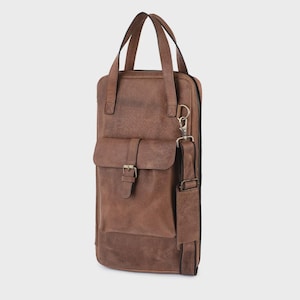 May include: A vertical brown bag with two top handles and a detachable shoulder strap. The bag features a front pocket with a buckle closure, a side zipper, and a metal D-ring attachment. The surface has a textured finish with visible stitching.