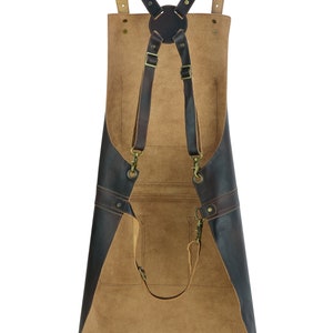 May include: A tan and dark brown leather apron featuring a cross-back strap design with adjustable metal buckles, brass-toned hardware, and multiple stitched pockets on the front surface. The apron has a smooth dark finish on the edges.