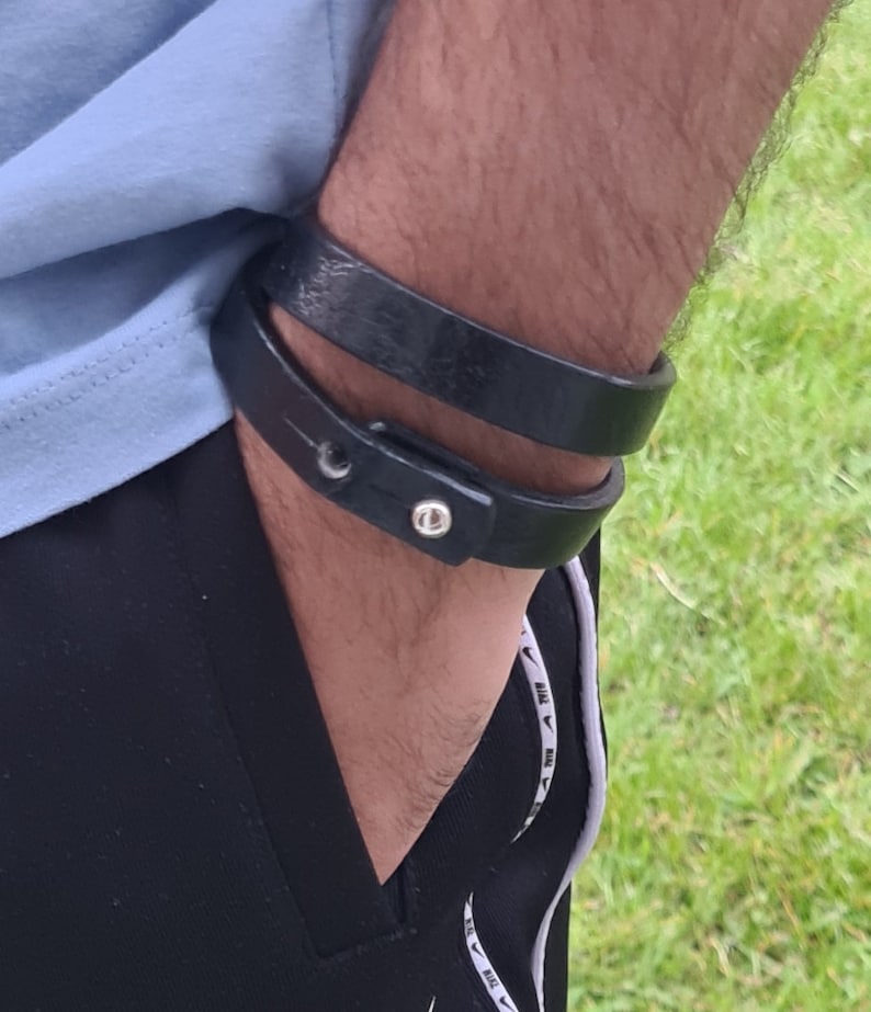 May include: A black leather bracelet with a silver buckle. The bracelet is worn on a person's wrist.