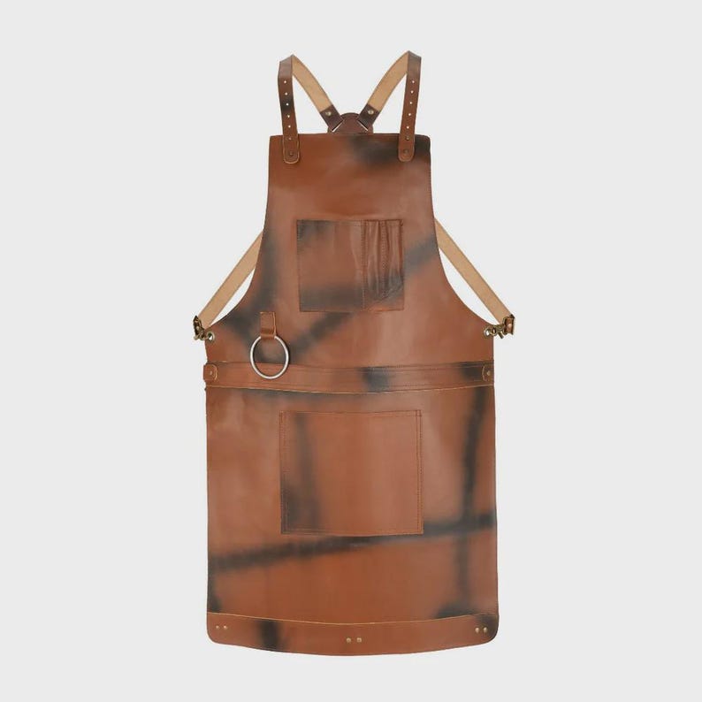 May include: A brown leather apron featuring adjustable shoulder straps with metal buckles, a chest pocket, a lower rectangular pocket, and a circular metal ring attached to the waist area. The surface has dark, distressed-style markings.