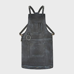 May include: A dark charcoal gray apron featuring adjustable shoulder straps with metal rivets, a chest pocket, a large lower pocket, and a circular metal ring attachment on the left side. The apron has a weathered finish with reinforced stitching.
