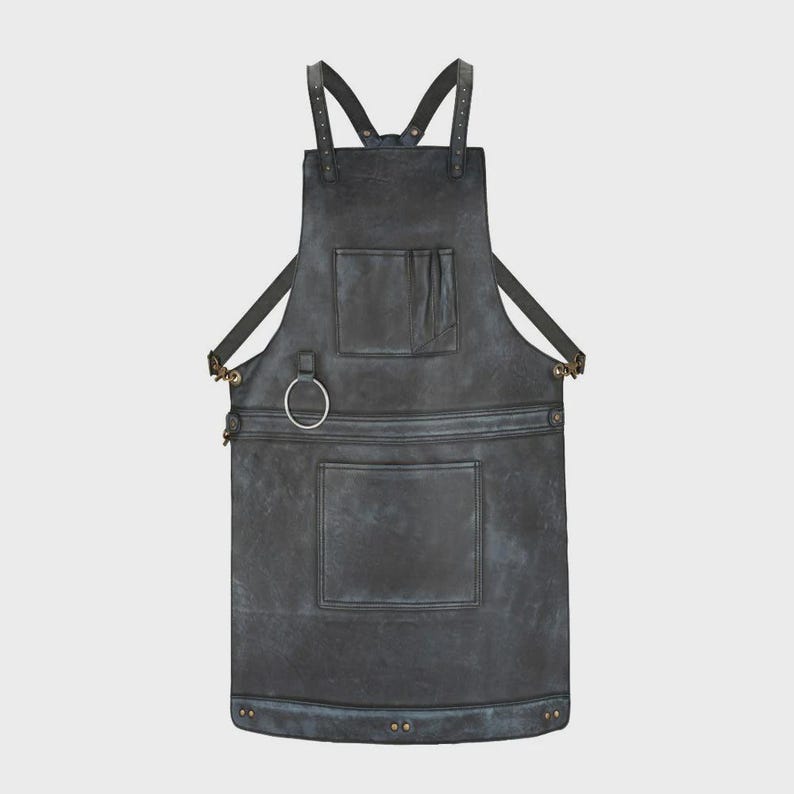 May include: A dark charcoal gray apron featuring adjustable shoulder straps with metal rivets, a chest pocket, a large lower pocket, and a circular metal ring attachment on the left side. The apron has a weathered finish with reinforced stitching.