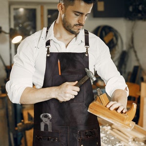May include: A dark brown leather apron featuring adjustable straps with metal hardware, a chest pocket containing a pencil, and multiple lower pockets holding a metal hammer and pliers. The logo LEATHERSHIRE is visible in the top left corner.
