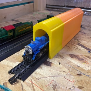 May include: A blue toy train engine is partially entering a yellow and orange plastic tunnel. The tunnel is on a wooden surface. The train is on a black track.