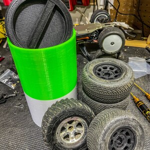 May include: A green and white cylindrical container with a black lid. The container is filled with four black tires with silver rims. The tires are stacked on top of each other.