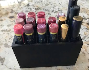 May include: A black rectangular lipstick organizer filled with various lipsticks. The lipsticks are in shades of pink, red, and nude, with some labeled "Pink", "Napa", and "Caramel". The organizer is on a light-colored countertop.