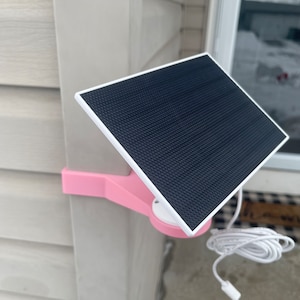 May include: A pink solar panel mount attached to a white siding wall. The solar panel is black and has a white border. A white cord is connected to the panel.