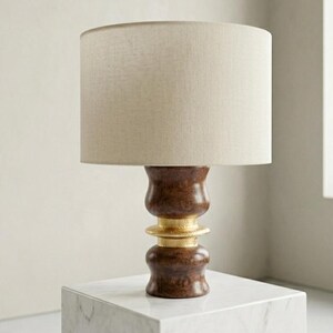 Può includere: Lampada da tavolo decorativa con paralume in tessuto beige. La base della lampada è in legno marrone scuro con dettagli dorati. La lampada è esposta su un cubo di marmo bianco.