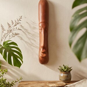 May include: A long, brown, wooden mask with carved facial features, including a nose, eyes, and mouth, hangs on a cream-colored wall. The mask is the focal point, with a natural wood grain finish. The image is a close-up of the mask.