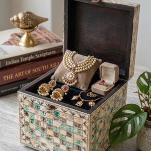 Può includere: Scatola portagioie in legno aperta con un motivo a scacchiera decorativo. All'interno, una collana, orecchini, un anello e altri gioielli in oro con perle e accenti rossi poggiano su una fodera di velluto nero.