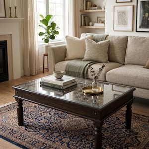 May include: A living room scene featuring a dark wood coffee table with a glass top, a beige sofa with decorative pillows, and a patterned rug. A plant sits near a window, and framed artwork hangs on the wall. The room has a warm, inviting atmosphere.