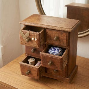 May include: A small, wooden jewelry chest with multiple drawers, some open to reveal contents. The chest is a warm brown color, with brass knobs. One drawer holds a gold butterfly pendant and pearl earrings. Another drawer contains a small, blue and white patterned box.