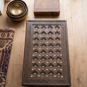 May include: A dark brown, rectangular coffee table with an ornate, carved surface and brass accents. The table is surrounded by a stack of brass bowls, a vintage book, and a patterned rug. The table is a focal point.
