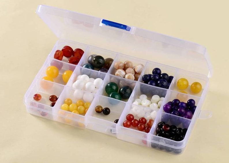 K&ouml;nnte beinhalten: Eine transparente Plastik-Aufbewahrungsbox mit 12 F&auml;chern, gef&uuml;llt mit bunten Perlen. Die Perlen sind in Reihen und Spalten angeordnet, wobei jedes Fach eine andere Farbe enth&auml;lt.