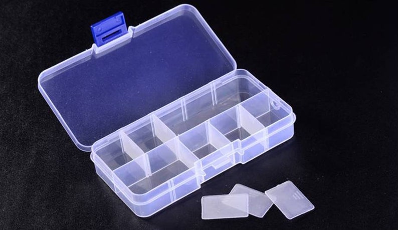K&ouml;nnte beinhalten: Eine transparente Plastik-Aufbewahrungsbox mit einem blauen Verschluss. Die Box hat sechs F&auml;cher und ist ge&ouml;ffnet, um die F&auml;cher zu zeigen. Neben der Box befinden sich f&uuml;nf transparente Plastik-Teiler auf der schwarzen Oberfl&auml;che.