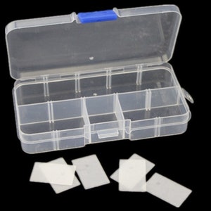 K&ouml;nnte beinhalten: Eine transparente Plastik-Aufbewahrungsbox mit einem blauen Verschluss. Die Box hat 6 F&auml;cher und ist ge&ouml;ffnet. Es befinden sich 6 kleine rechteckige wei&szlig;e Teile auf einem schwarzen Hintergrund.