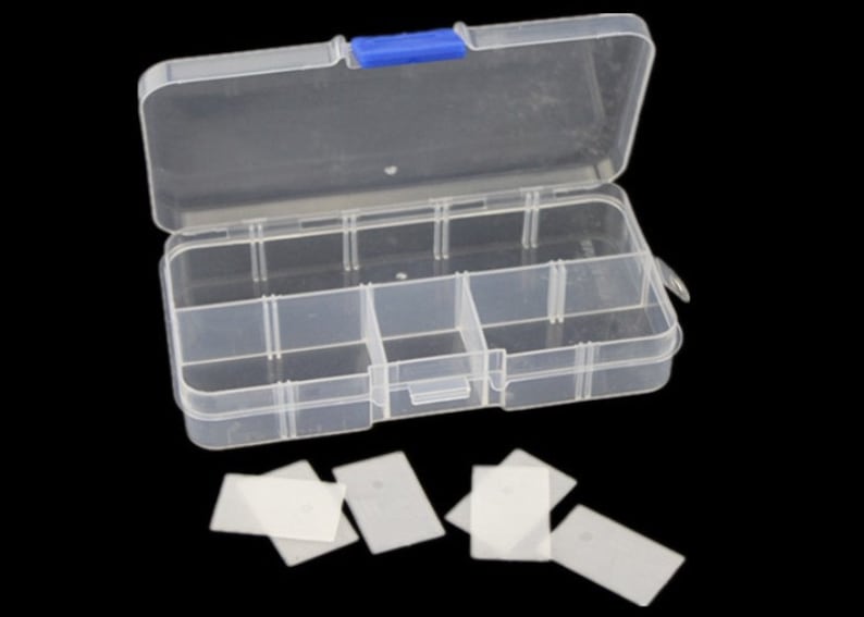 K&ouml;nnte beinhalten: Eine transparente Plastik-Aufbewahrungsbox mit einem blauen Verschluss. Die Box hat 6 F&auml;cher und ist ge&ouml;ffnet. Es befinden sich 6 kleine rechteckige wei&szlig;e Teile auf einem schwarzen Hintergrund.