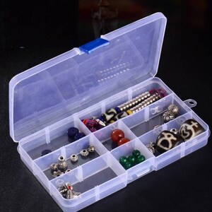 K&ouml;nnte beinhalten: Eine transparente Plastik-Aufbewahrungsbox mit einem blauen Verschluss. Die Box hat acht F&auml;cher und ist mit verschiedenen Perlen, Charms und Schmuck gef&uuml;llt.