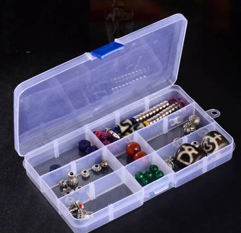 K&ouml;nnte beinhalten: Eine transparente Plastik-Aufbewahrungsbox mit einem blauen Verschluss. Die Box hat acht F&auml;cher und ist mit verschiedenen Perlen, Charms und Schmuck gef&uuml;llt.