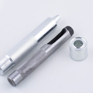 May include: Two silver metal tools for punching holes in leather, one with a black ring, and a silver metal grommet.