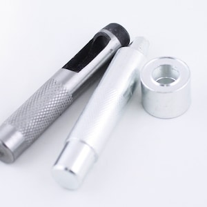 May include: A set of silver leather hole punch tools. The set includes a punch, a hammer, and a grommet. The punch has a black handle and a silver metal head. The hammer has a silver metal head and a silver metal handle. The grommet is silver metal.