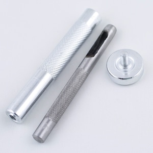 May include: A set of three silver-toned tools for leather crafting. The tools include a punch, a hammer, and a base. The punch has a sharp, pointed end and a textured handle. The hammer has a round head and a textured handle. The base is a flat, round disc with a raised center.
