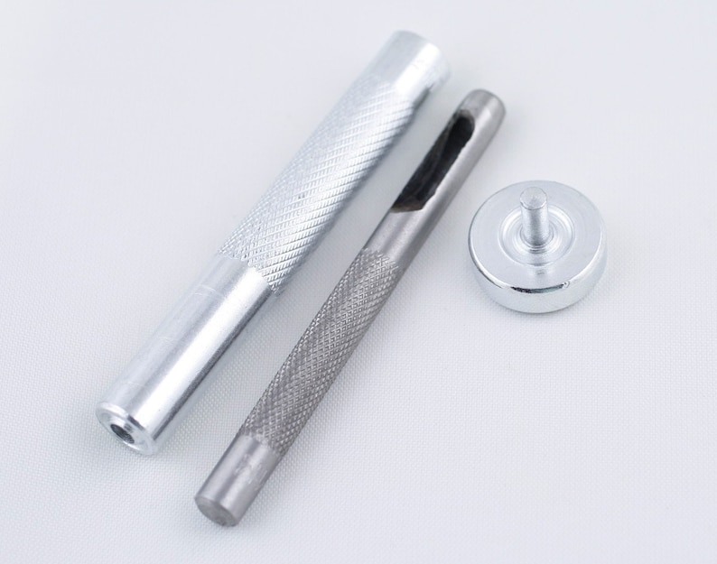 May include: A set of three silver-toned tools for leather crafting. The tools include a punch, a hammer, and a base. The punch has a sharp, pointed end and a textured handle. The hammer has a round head and a textured handle. The base is a flat, round disc with a raised center.