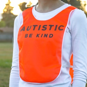 Puede incluir: Chaleco de malla naranja brillante con las palabras "AUTISTIC BE KIND" impresas en negro. El chaleco se lleva sobre una camisa blanca de manga larga. El fondo muestra un campo verde y una porter&iacute;a de f&uacute;tbol.