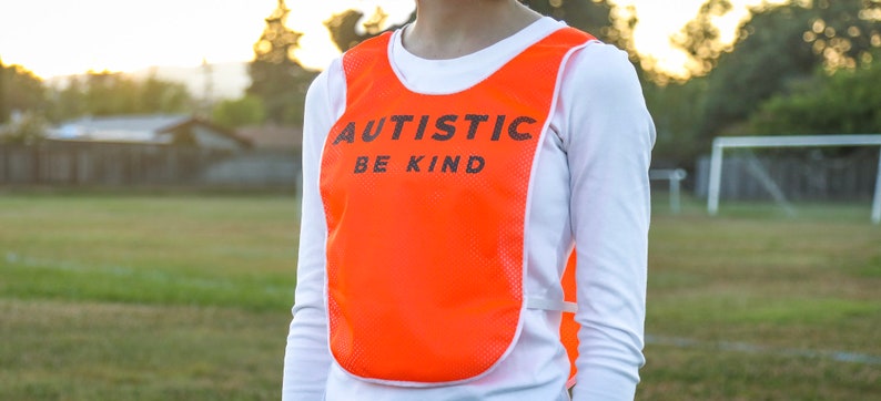 Puede incluir: Chaleco de malla naranja brillante con las palabras "AUTISTIC BE KIND" impresas en negro. El chaleco se lleva sobre una camisa blanca de manga larga. El fondo muestra un campo verde y una porter&iacute;a de f&uacute;tbol.