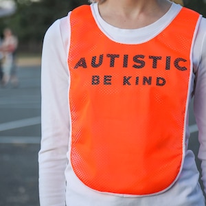 Puede incluir: Un chaleco de malla naranja con ribete blanco, con el texto "AUTISTIC BE KIND" en letras negras. El chaleco se lleva sobre una camiseta blanca de manga larga. El fondo muestra una escena borrosa al aire libre.