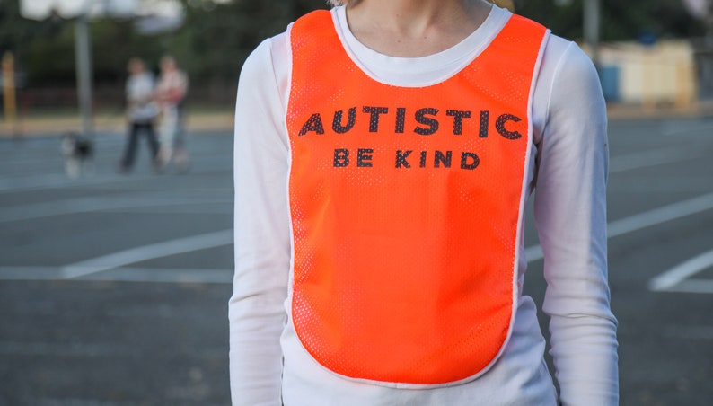 Puede incluir: Un chaleco de malla naranja con ribete blanco, con el texto "AUTISTIC BE KIND" en letras negras. El chaleco se lleva sobre una camiseta blanca de manga larga. El fondo muestra una escena borrosa al aire libre.
