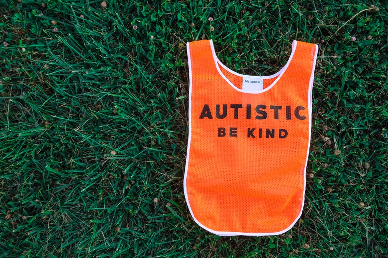 Puede incluir: Un chaleco de malla naranja con ribete blanco y las palabras "AUTISTIC BE KIND" en negro. El chaleco est&aacute; sobre hierba verde. La parte superior del chaleco tiene una peque&ntilde;a etiqueta blanca que dice "My name is:"