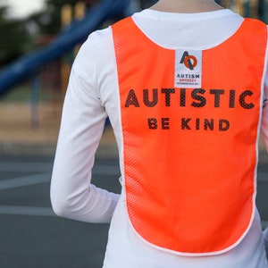 Puede incluir: Un chaleco de malla naranja con las palabras "AUTISTIC BE KIND" impresas en negro. El chaleco tambi&eacute;n presenta un logotipo de "AUTISM ODYSSEY". El chaleco se lleva sobre una camiseta blanca de manga larga.