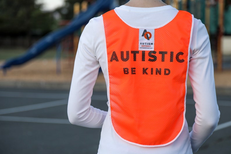 Puede incluir: Un chaleco de malla naranja con las palabras "AUTISTIC BE KIND" impresas en negro. El chaleco tambi&eacute;n presenta un logotipo de "AUTISM ODYSSEY". El chaleco se lleva sobre una camiseta blanca de manga larga.