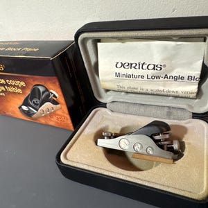 May include: A Veritas miniature low-angle block plane in a black case with a tan interior. The box next to it has the text "Veritas" and "Rabot de coupe à angle faible miniature". The plane is silver and black.