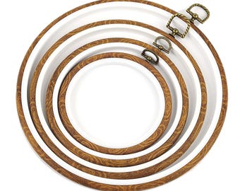May include: A set of four circular embroidery hoops nested inside one another. Each hoop features a brown wood-grain pattern on the outer ring and a smooth white inner ring. Each hoop includes a small, antique brass-colored metal tightening screw.