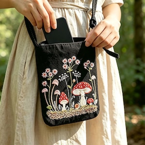 Puede incluir: Un bolso bandolera negro con bordados detallados de varios hongos y flores silvestres en tonos rojos, blancos y verdes. El bolso rectangular cuenta con un cierre de cremallera y una correa negra fina con un diseño botánico frontal.