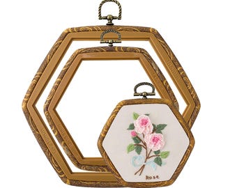 May include: Three nested hexagonal embroidery hoops with a wood-grain finish and bronze-toned hardware. The smallest hoop contains a white fabric panel featuring an embroidered pink rose bouquet with green leaves, a light blue ribbon, and the text Rose.