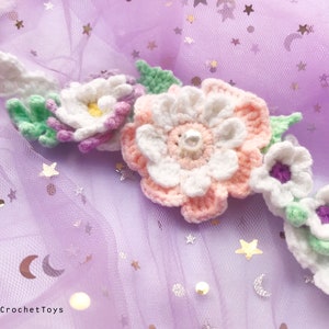 May include: A crocheted flower headband with white, pink, and purple flowers and green leaves. The headband is on a purple background with silver stars.