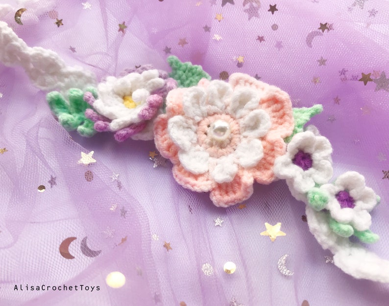 May include: A crocheted flower headband with white, pink, and purple flowers and green leaves. The headband is on a purple background with silver stars.